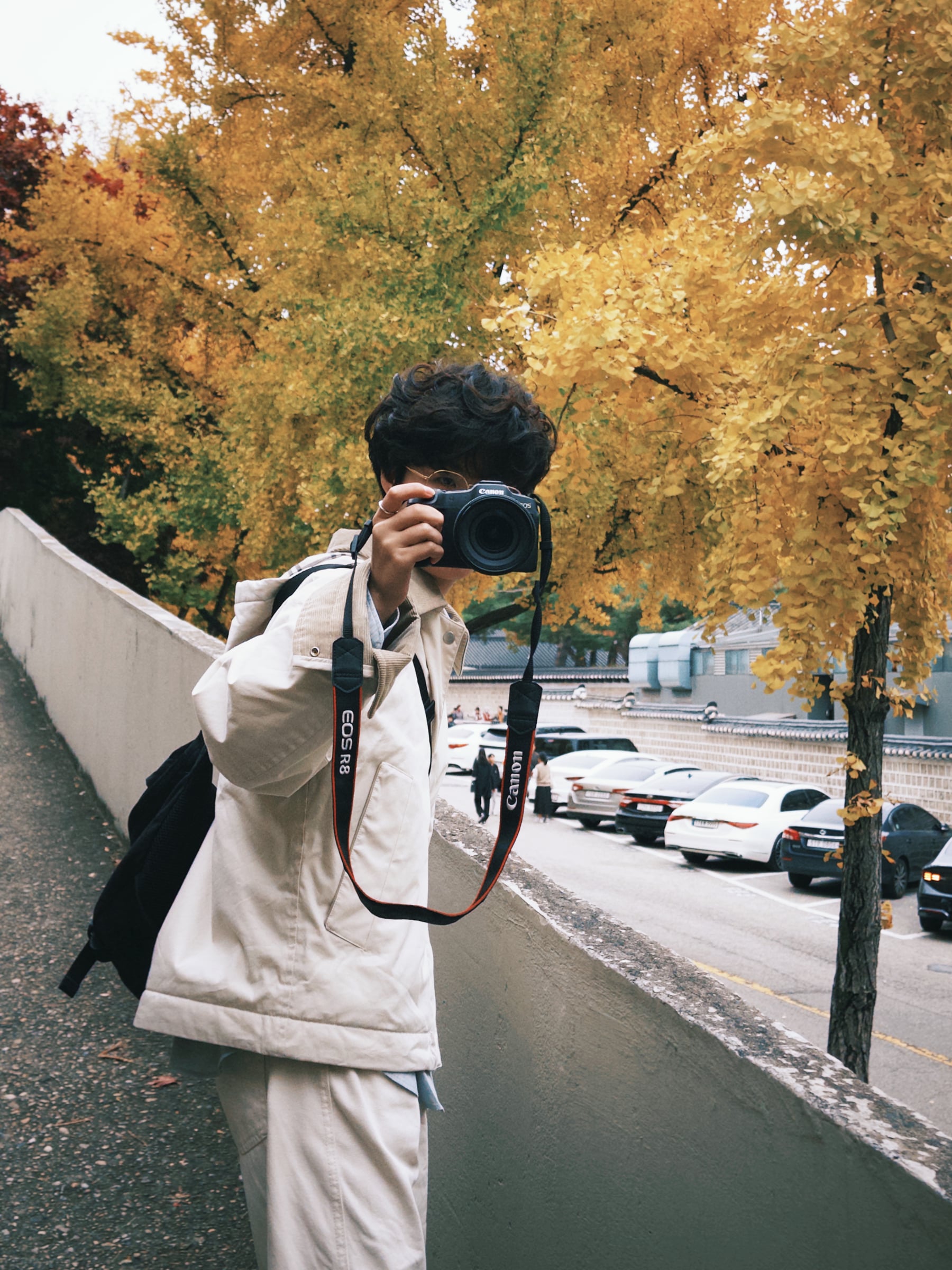 Rayce Zhang holding a camera beneath yellow autumn leaves.